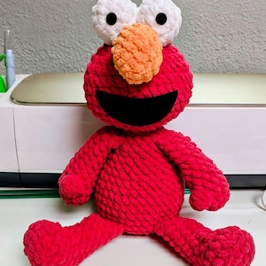 May include: A handmade red plush toy with white eyes, a bright orange nose, and a large black smile. The plush toy is sitting with its legs spread out. The toy is made of a soft, textured material.