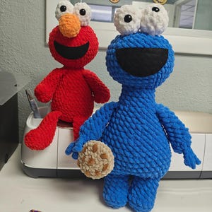 May include: Two handmade crochet plush toys: a red character with an orange nose and a blue character holding a cookie. A crochet hook with a patterned handle rests on the surface. The toys are on a white surface.