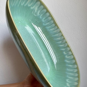 Peut inclure: Un grand bol en céramique de forme ovale présentant un intérieur vert menthe clair brillant avec un motif distinctif en forme de plumes ou de coups de pinceau rayonnant du centre vers le bord. Le bord extérieur est fini par une bordure jaune pâle.