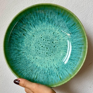 Puede incluir: Un cuenco de cerámica redondo con un esmalte vibrante en tonos turquesa y verde azulado, con un patrón radiante y moteado que se funde en un borde verde sólido. La superficie interior muestra un efecto texturizado con tonos azules.