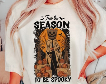 May include: A cream-coloured short-sleeve t-shirt featuring a graphic print with the text "This the SEASON TO BE SPOOKY". The design depicts a scarecrow figure with a pumpkin head holding a pitchfork, surrounded by pumpkins and corn stalks.