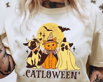 Könnte beinhalten: Ein cremefarbenes T-Shirt mit einer Grafik von drei Katzen in Halloween-Kostümen, darunter ein Teufel, eine Hexe und ein Geist, vor einem gelben Mond mit Fledermäusen. Der Schriftzug CATLOWEEN ist in dunkelbraunen Buchstaben unter den Katzen gedruckt.