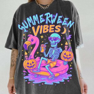 May include: Dark gray t-shirt with a colorful graphic that reads "Summerween Vibes." The graphic features a skeleton wearing sunglasses, sitting on a flamingo float, and holding a drink. Pumpkins and candles are also in the design.