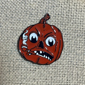 May include: A red enamel pin in the shape of a pumpkin with a cartoon face, smoking a cigarette. The pin has a black outline and white eyes and teeth. The pin is on a tan fabric background.