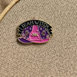 May include: Enamel pin featuring a witch hat in shades of pink and purple, with the words "TEAM WITCH" on a black banner. The pin includes hand and moon symbols, and is set against a neutral fabric background.