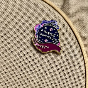 May include: A gold-rimmed enamel pin featuring a dark purple hand holding a navy blue crystal ball. The crystal ball contains pink stars and the white text MAKE YOUR MAGIC, surrounded by swirling purple and pink smoke-like accents.