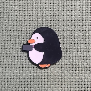 May include: A penguin-shaped enamel pin with a black body, light pink belly, orange feet and beak, and a small black rectangle. The pin is set against a gray textured background.