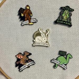 May include: A collection of enamel pins depicting mythical creatures. The pins include Bigfoot, aliens, a jackalope, the Jersey Devil, and Nessie. Each pin features the text "BELIEVES IN YOU".