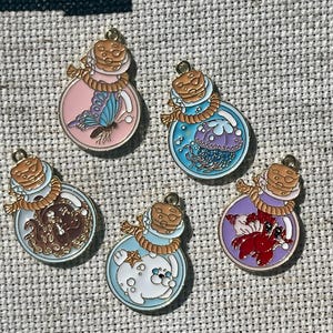May include: Six gold-colored charms shaped like glass bottles with cork tops. Each bottle contains a different creature: a butterfly, a jellyfish, an octopus, a dog, a crab, and a starfish. The charms are on a beige fabric background.