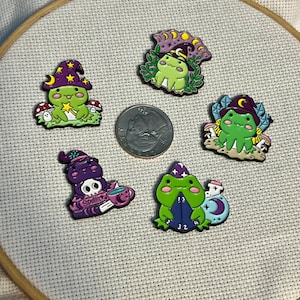 May include: Five colorful enamel pins featuring whimsical frog designs. Each frog is depicted in a wizard or witch theme, wearing hats and surrounded by magical elements like mushrooms, moons, and spell books. The pins are set on a white fabric background.