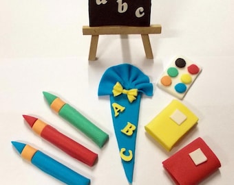 May include: A set of school-themed cake toppers, including a blue and yellow cone with the letters ABC, a small easel with a black chalkboard that says 'abc', three crayons in red, blue, and green, a yellow and red book, a red book, and a paint palette with six colorful paint dots.