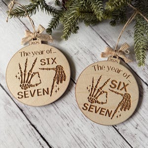 May include: Two wooden Christmas ornaments with the text "The year of... SIX SEVEN" and a skeleton hand design. Each ornament has a twine hanger with a small bow and the year "2025" printed on it. The ornaments are displayed on a wooden surface.
