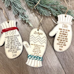 Puede incluir: Tres adornos navideños de madera con forma de mitón con colgadores de cuerda. Cada adorno tiene un nombre, un poema y el año 2025 grabados. Los mitones están decorados con hilo rojo, verde y blanco.