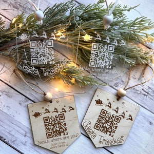 Op de afbeelding: Drie kerstversieringen met een Santa Tracker-thema. Twee zijn gemaakt van helder acryl met een wit touwtje en een witte kraal. De derde is gemaakt van hout met een bruin touwtje en een bruine kraal. Elke versiering heeft een QR-code en de tekst "Santa Tracker" en "Santa, waar ben je? Scan om te zien waar Santa is!"