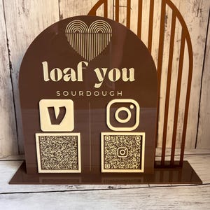 Puede incluir: Un letrero de acrílico marrón con un diseño de corazón y el texto "loaf you sourdough". El letrero tiene dos códigos QR, uno para Instagram y otro para Venmo.