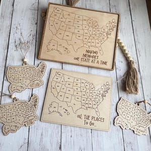 Könnte beinhalten: Holzkarte der Vereinigten Staaten mit Umrissen der Bundesstaaten und Text. Zwei rechteckige Tafeln und drei Ornamente sind sichtbar. Der Text auf den Tafeln lautet "MAKING MEMORIES ONE STATE AT A TIME" und "OH, THE PLACES TO GO..."