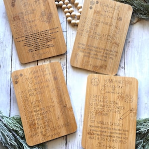 Puede incluir: Cuatro tablas de recetas de madera con instrucciones grabadas para hacer peanut brittle, galletas de jengibre, chocolate caliente y galletas de azúcar. Cada tabla presenta un diseño festivo y la frase "Enjoy!"