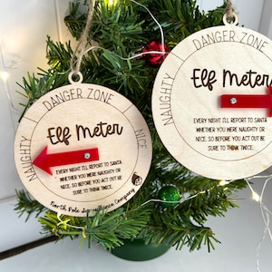 Puede incluir: Dos adornos navideños de madera con el texto "Elf Meter" y una flecha roja. Los adornos tienen las frases "Danger Zone", "Naughty" y "Nice". El texto dice: "Cada noche informaré a Santa si fuiste travieso o bueno."