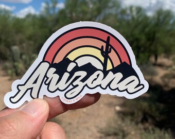 May include: A sticker with a retro sunset design featuring a silhouette of mountains and a cactus. The text "Arizona" is written in a stylized font with a vintage aesthetic. The sticker has a white border and the text "Stray x Wild" is printed in small letters at the bottom.