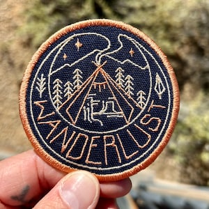 May include: A round, navy blue embroidered patch with a tan outline. The patch features a stylized image of a tent with smoke rising from the top, surrounded by trees and stars. The text "WANDERLUST" is written in a circular pattern around the image.
