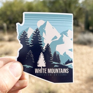 May include: A blue and white sticker featuring a stylized illustration of the White Mountains in Northern Arizona. The sticker depicts a mountain range with a moon in the sky and pine trees in the foreground. The text "White Mountains" and "Northern Arizona" is printed on the sticker.