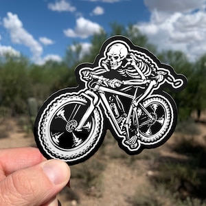 May include: Black and white sticker of a skeleton riding a mountain bike. The skeleton is wearing a helmet and has a determined expression on its face. The bike has knobby tires and a suspension fork.