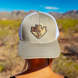 May include: A gray and white trucker hat with a brown and gold embroidered patch of a cactus and a sun. The patch is in the shape of a heart.