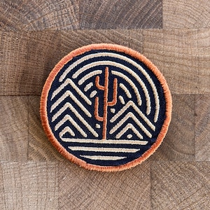 May include: Circular embroidered patch featuring a desert landscape design. The patch has a navy blue background with a cream-colored mountain range and concentric lines. A rust-colored cactus is in the center, and the patch is bordered with orange thread.