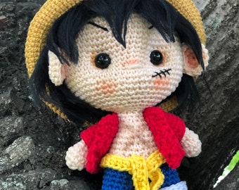 May include: Crocheted amigurumi doll with black hair, a yellow straw hat, a red shirt, blue pants, and a yellow belt. The doll has a stitched-on scar on its face.