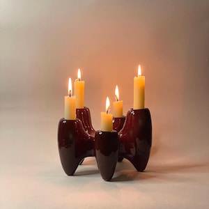 May include: A dark red, four-armed ceramic candle holder with a glossy finish. The object features four distinct cylindrical sockets, each holding a lit, cream-coloured taper candle with a visible flame, creating a warm, glowing light effect.
