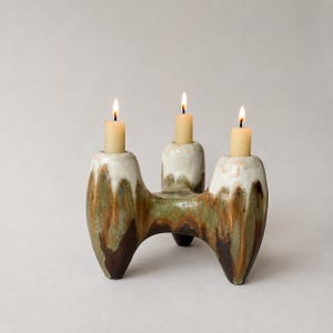 Könnte beinhalten: Ein Kerzenständer aus Keramik mit drei brennenden Stabkerzen. Die Basis ist mit einer Glasur in tropfenden Mustern in Creme, Salbeigrün und Dunkelbraun versehen. Die Struktur hat drei zylindrische Halterungen und eine geschwungene Basis.