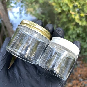 Peut inclure: Deux bocaux en verre transparent avec des couvercles. Le plus grand bocal a un couvercle doré et le plus petit bocal a un couvercle blanc.