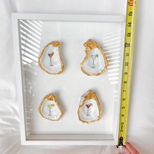 May include: A framed artwork featuring four oyster shells, each with a gold-painted rim and a cocktail illustration. The frame is white. The artwork is approximately 10 inches wide.