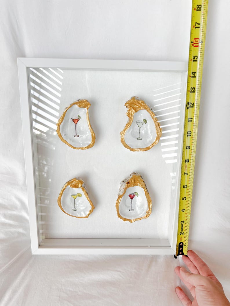 May include: A framed artwork featuring four oyster shells, each with a gold-painted rim and a cocktail illustration. The frame is white. The artwork is approximately 10 inches wide.