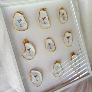 May include: A framed artwork featuring nine oyster shells, each painted with a different cocktail design. The shells have gold-painted edges and are arranged on a white background within a white frame. The cocktails are in various colors.