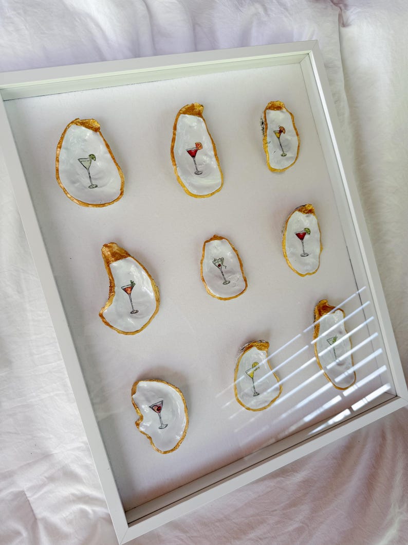 May include: A framed artwork featuring nine oyster shells, each painted with a different cocktail design. The shells have gold-painted edges and are arranged on a white background within a white frame. The cocktails are in various colors.