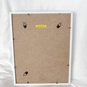 May include: Back of a white framed picture with a brown backing. The frame has four metal hanging brackets and a yellow sticker that reads "Do Not Hang With Wire".