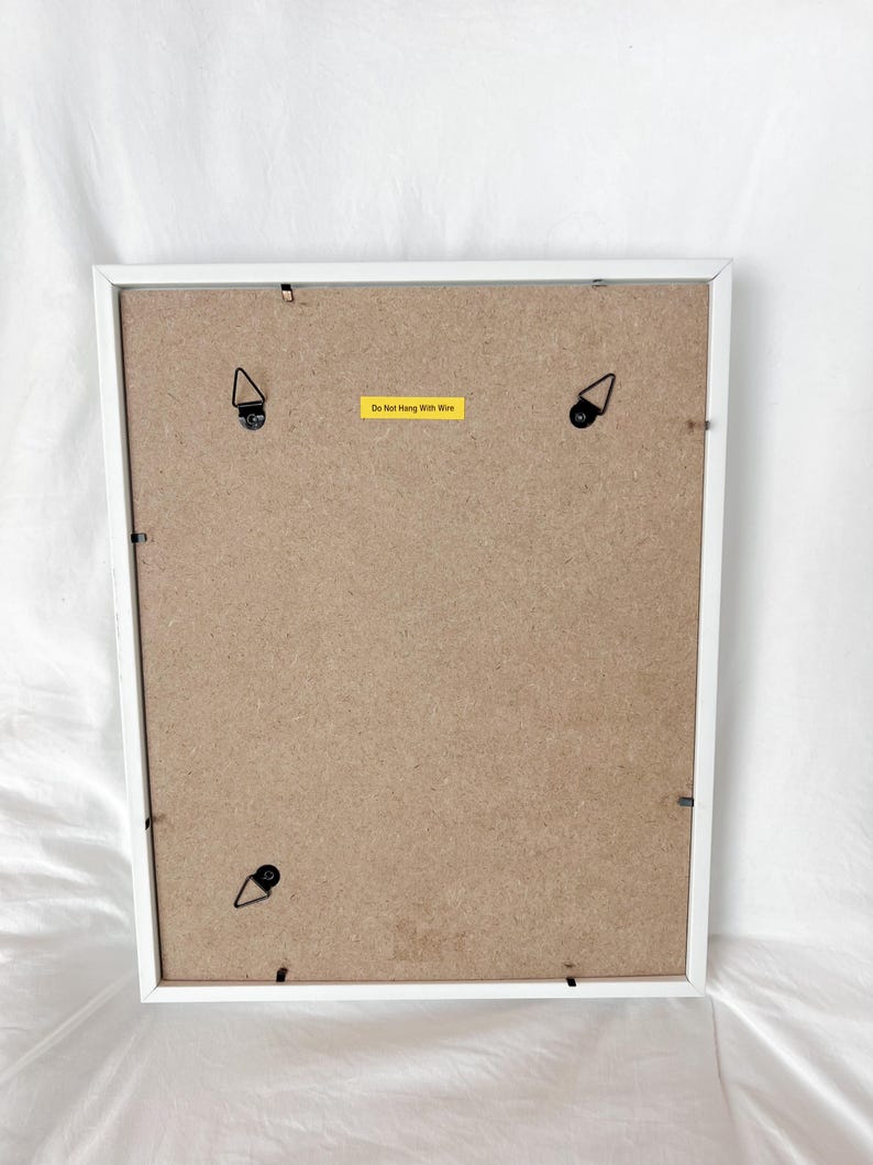 May include: Back of a white framed picture with a brown backing. The frame has four metal hanging brackets and a yellow sticker that reads "Do Not Hang With Wire".
