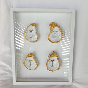 Può includere: Opera incorniciata con quattro conchiglie di ostrica con bordi dorati, ognuna con un'illustrazione di cocktail in rosso, verde e giallo. La cornice bianca completa il tema costiero.