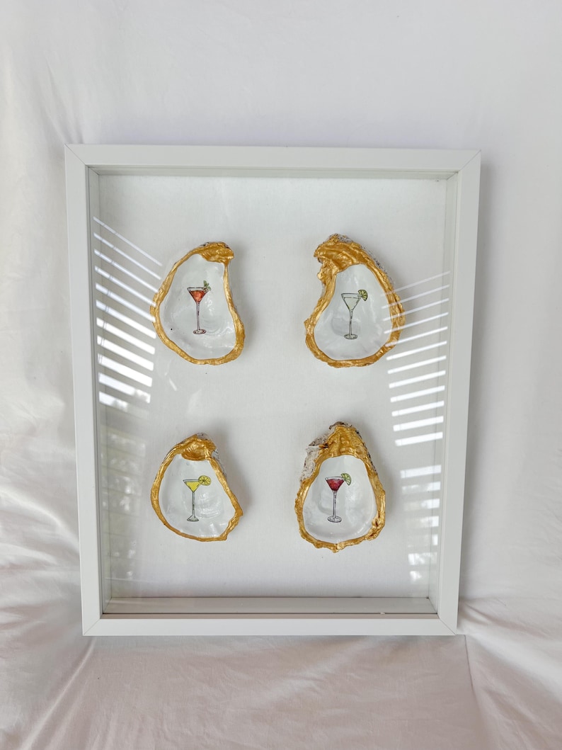 May include: A framed artwork featuring four oyster shells with gold-painted edges. Each shell holds a miniature cocktail illustration in red, green, and yellow. The white frame complements the artwork's coastal theme.