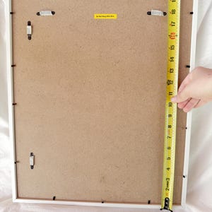 May include: Back of a white framed picture. A yellow measuring tape shows the frame is approximately 21 inches tall. The back is a light brown color with hanging hardware and a yellow sticker that reads "Do Not Hang With Wire."