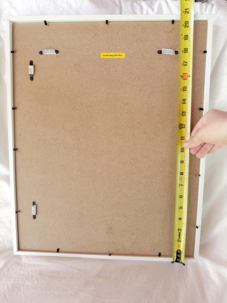 May include: Back of a white framed picture. A yellow measuring tape shows the frame is approximately 21 inches tall. The back is a light brown color with hanging hardware and a yellow sticker that reads "Do Not Hang With Wire."
