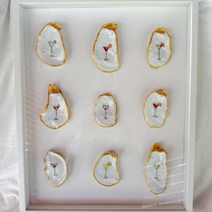 May include: A framed artwork featuring eleven oyster shells, each painted with a cocktail design. The shells have a white interior and gold edging. The cocktails are in various colors, including red, yellow, and green.