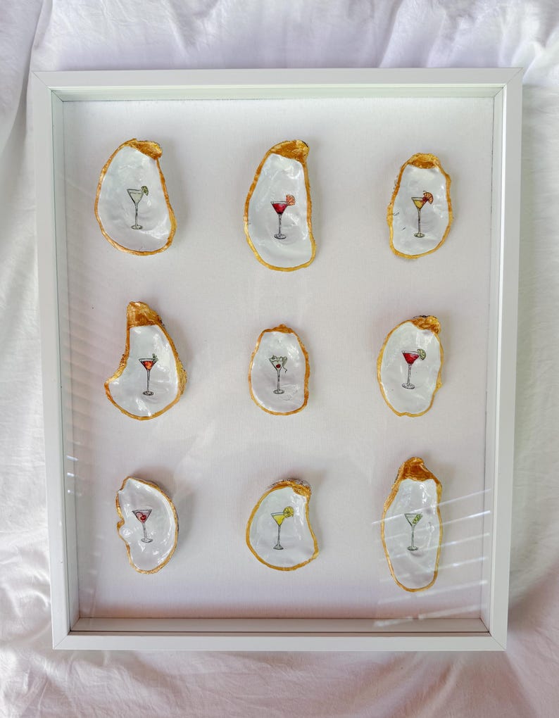 May include: A framed artwork featuring eleven oyster shells, each painted with a cocktail design. The shells have a white interior and gold edging. The cocktails are in various colors, including red, yellow, and green.
