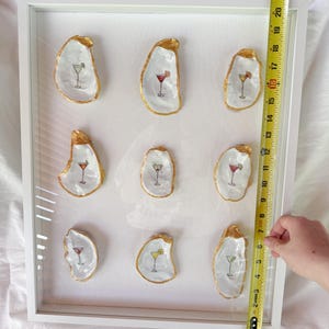 May include: A framed artwork featuring nine oyster shells, each with a gold-painted edge and a cocktail illustration. The cocktails are depicted in various colors. The frame is white, and the artwork is approximately 18 inches tall.