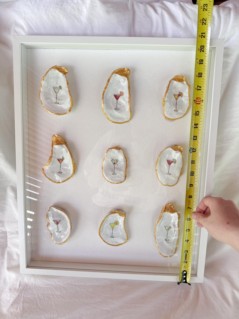 May include: A framed artwork featuring nine oyster shells, each with a gold-painted edge and a cocktail illustration. The cocktails are depicted in various colors. The frame is white, and the artwork is approximately 18 inches tall.