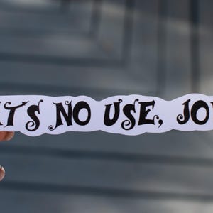May include: A white sticker with the phrase "IT'S NO USE, JO!" in black, stylized text. The sticker has a cloud-like shape and is held against a blurred background. The text is in a bold, decorative font.