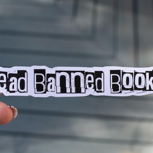 May include: A white sticker with the words "Read Banned Books!" in a black, cut-out style font. The sticker is held by a person with manicured nails. The background is a blurred, gray surface.
