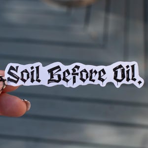 以下が含まれることがあります： ゴシック体の「Soil Before Oil」というフレーズが書かれた白いステッカー。ステッカーは灰色の背景に対して保持されています。テキストは黒色です。