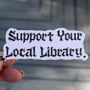 May include: A white sticker with the text "Support Your Local Library" in a gothic font. The sticker is held up against a blurred background. The sticker is a rectangle with rounded edges.